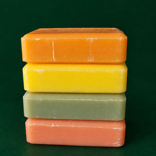 Four rectangular bars of soap stacked vertically on a dark green background. From top to bottom, the soap colors are orange, yellow, sage green, and coral pink.