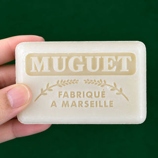A hand holding a rectangular bar of MUGUET Fabrique A Marseille soap against a solid dark green background.