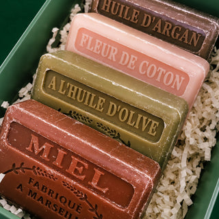 Four artisanal soap bars in a green gift box with shredded paper filler. The soaps are labeled in French with their respective oils: Miel (beige), A L'HUILE D'OLIVE (green), Fleur de Coton (pink), and Huile d'Argan (dark brown).