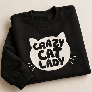 A folded black crewneck sweatshirt with a white graphic of a cat head and the text 'CRAZY CAT LADY' printed on the front, displayed on a light beige background.