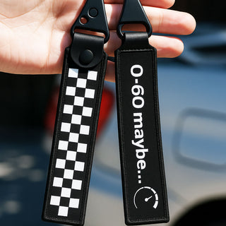 Two black faux-leather keychain tags held in a hand. One tag has a black and white checkered pattern, and the other has white text reading '0-60 maybe.