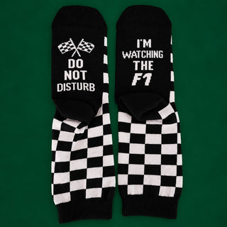 A pair of black crew socks with white checkered patterns and text, displayed flat against a solid green background. One sock features crossed checkered flags with the text 'DO NOT DISTURB', and the other features the text 'I'M WATCHING THE F1'.