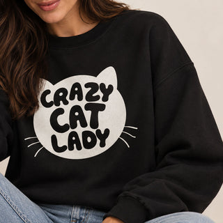 A person wearing a black crewneck sweatshirt with a large white cat head graphic and the words 'CRAZY CAT LADY' in black lettering on the front, paired with blue jeans against a light neutral background.