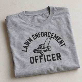 A folded heather gray t-shirt with a black graphic print that reads 'LAWN ENFORCEMENT OFFICER' around a lawn mower illustration, displayed on a light beige textured background.