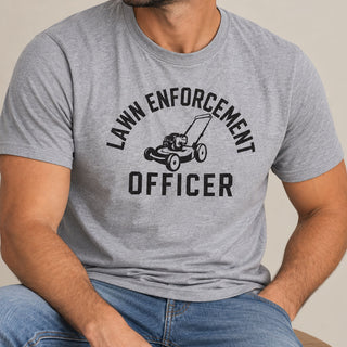 A man wearing a heather gray crewneck t-shirt with a black graphic that reads 'LAWN ENFORCEMENT OFFICER' around a lawnmower illustration, paired with blue jeans.