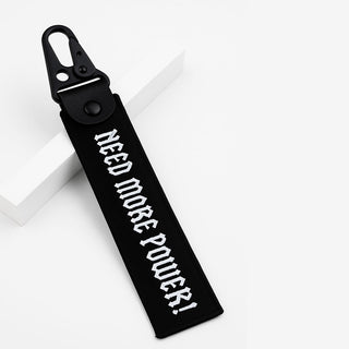 A black fabric keychain or bag tag with a black metal clip, featuring the text 'NEED MORE POWER!' in white gothic-style lettering, displayed on a clean white background.