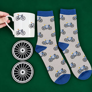 A pair of crew socks with a bicycle pattern on a light gray background and blue cuffs, heels, and toes, displayed next to a white ceramic mug with the same bicycle print and two matching coaster lids on a dark green background.