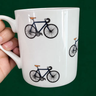 A white ceramic mug with a handle, decorated with illustrations of navy blue bicycles that have orange seats, held by a hand against a solid dark green background.