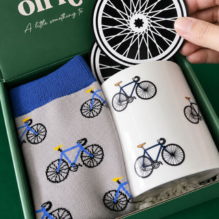 A bicycle-themed gift set in a green box, featuring a pair of gray socks with blue and black bicycle prints and a white ceramic mug with matching bicycle illustrations. A hand is holding a black and white coaster with a bicycle wheel design.