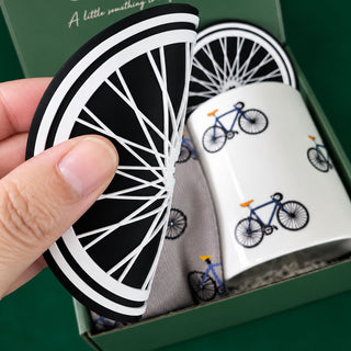 A hand holds a black and white bicycle-themed coaster in front of a white ceramic mug printed with blue bicycles, inside a green gift box with matching coasters and a gray bicycle-patterned cloth.