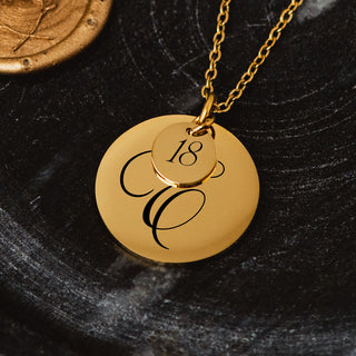 Gold personalised disc necklace with engraved script initial and small “18” charm on a chain, displayed on a dark textured surface.