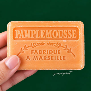 A hand holding a rectangular orange soap bar embossed with 'PAMPLEMOUSSE FABRIQUÉ A MARSEILLE' against a dark green background.