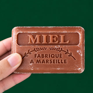 A hand holding a rectangular brown soap bar embossed with 'MIEL FABRIQUE A MARSEILLE' against a solid dark green background.