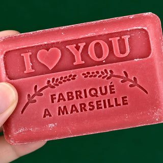A close-up of a hand holding a pink rectangular bar of soap embossed with 'I ❤️ YOU' and 'FABRIQUÉ A MARSEILLE' against a dark green background.