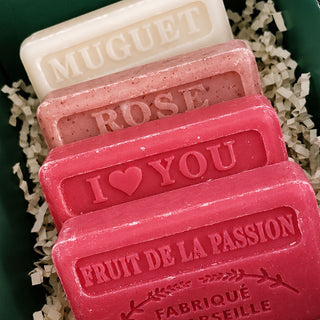 Four decorative handmade soap bars arranged in a stack on shredded paper inside a dark green box. The soaps are embossed with names and phrases: 'MUGuet', 'ROSE', 'I <heart> YOU', and 'FRUIT DE LA PASSION FABRIQUE PAR SEILLE'.