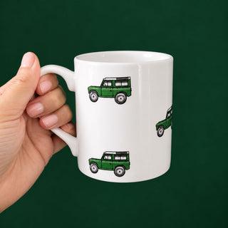 A hand holding a white ceramic mug with a repeating pattern of a land rover set against a solid dark green background.