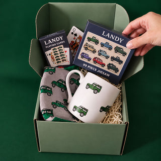A curated gift box set containing a 99-piece jigsaw puzzle, a deck of Landy playing cards, a mug, and a socks, all featuring green vintage military vehicles. A hand is holding the puzzle box, which is displayed inside an open sage green cardboard box on a solid dark green background.