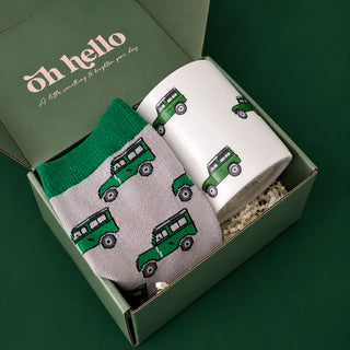 A gift set in a sage green box featuring a light gray knit sock with a green jeep pattern and a white ceramic mug with matching green jeeps, set against a dark green background.