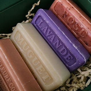 Four embossed soap bars in peach, cream, purple, and coral pink, arranged in shredded paper inside a dark green box. The soaps are labeled with scents including MUGUET, LAVANDE, and ROSE.
