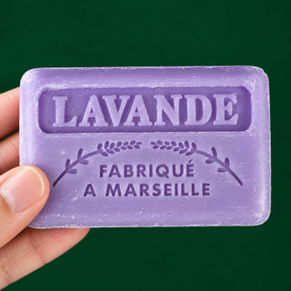 A hand holding a lavender-colored rectangular bar of soap embossed with the text 'LAVANDE FABRIQUÉ A MARSEILLE' against a solid dark green background.