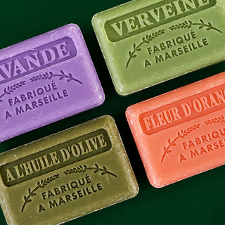 Four rectangular bars of Marseille soap arranged in a flat-lay pattern on a dark green background. The soaps are in four different colors: purple (lavande), light green (verveine), olive green (a l'huile d'olive), and coral orange (fleur d'orange).