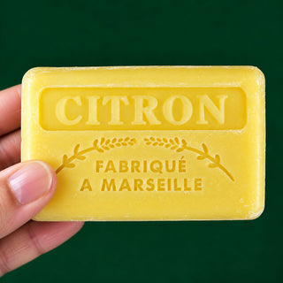 A hand holding a rectangular yellow citron soap bar embossed with 'CITRON FABRIQUÉ A MARSEILLE' against a solid dark green background.