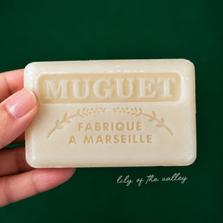 A hand holding a rectangular bar of MUGUET Fabrique à Marseille soap against a solid dark green background.