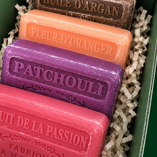 Four rectangular handmade soaps in different colors arranged in a green box with white crinkle paper filler. The soaps are labeled in French with 'PATCHOULI', 'FLEUR D'ORANGER', 'ROSE D'ARGAN', and 'FRUIT DE LA PASSION'.
