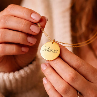 Gold personalised disc necklace engraved with Mama, held in hands on a soft neutral background