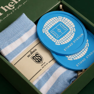 A gift set in a dark green box containing light blue and white striped socks with a paper band reading 'SOLE SUPPORTER' and two bright blue circular coasters printed with a white stadium and 'ETIHAD STADIUM' text.