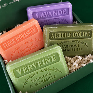 Four artisanal bar soaps in different colors (purple lavender, orange fleur d'oranger, and two green scents) arranged in a green gift box with shredded paper filler. Each soap is embossed with its respective French scent name and 'FABRIQUÉ A MARSEILLE'.
