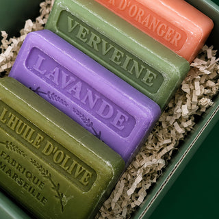 Four rectangular soap bars in olive green, purple, peach, and dark green, arranged in shredded paper inside a green box. The soaps are embossed with their names: 'LAVANDE', 'VERVEINE', and 'D'ORANGER', plus 'L'HUILE D'OLIVE' and 'FABRIQUE MARSEILLaise' on the front soap.