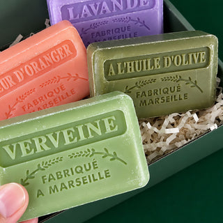Four Marseille-style bar soaps in different colors are arranged in a green gift box with shredded paper. A hand is holding the green soap in the foreground.