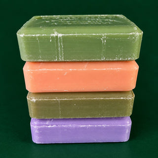 Four rectangular bars of soap stacked vertically on a dark green background. The top bar is olive green, followed by peach, olive green, and lavender purple bars.