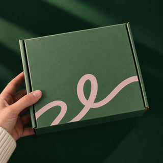 A hand holding a dark green square box with a light pink abstract line design on the lid, set against a matching dark green background.