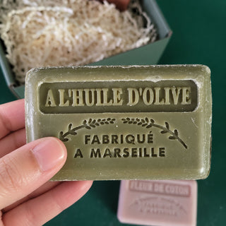 A hand holds a rectangular olive-green bar of soap embossed with 'A L'HUILE D'OLIVE FABRIQUE A MARSEILLE'. In the background, a green box filled with shredded paper is visible, along with a small pink soap bar labeled 'FLEUR DE CITRON'.