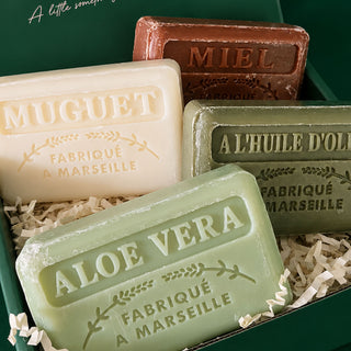 Four embossed bars of Marseille soap in a green gift box with shredded paper filler. The soaps are labeled ALOE VERA (light green), MUGUET (cream), A L'HUILE D'OLIVE (olive green), and MIEL (brown).