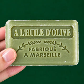 A hand holding a rectangular bar of olive oil soap in a muted olive green color, featuring embossed French text 'A L'HUILE D'OLIVE' and 'FABRIQUÉ A MARSEILLE' against a solid dark green background.