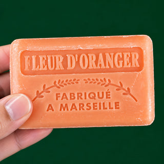 A hand holding a rectangular orange soap bar embossed with the text 'FLEUR D'ORANGER' and 'FABRIQUÉ A MARSEILLE' against a solid dark green background.