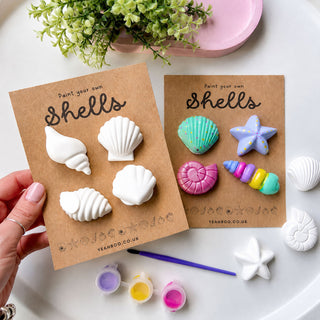 Two brown cardboard cards displaying painted seashell and starfish craft designs, held by a hand over a white surface with small paint pots, a paintbrush, and a pink dish in the background.