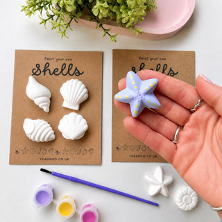 A hand holds a painted lavender starfish shell charm with yellow dots, shown next to two brown cards displaying unpainted white seashell charms and a paintbrush, on a white background with a pink tray and green plant in the background.