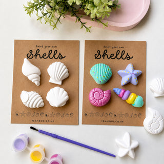 Two brown cards labeled 'Paint your own Shells' with unpainted white shell-shaped items, displayed on a light surface with paint pots, a paintbrush, a starfish, and greenery. One side shows painted shell designs in turquoise, pink, purple, yellow, and blue.