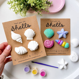 A hand holds a card displaying white shell-shaped craft items, with another card showing colorful painted shell designs. The scene includes small paint pots, a paintbrush, loose shell-shaped pieces, and a plant on a white surface.