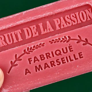 A close-up of a pink soap bar embossed with the French text 'FRUIT DE LA PASSION' and 'FABRIQUE A MARSEILLE', held by a hand against a dark green background.