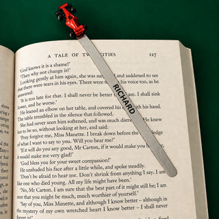A silver metal bookmark with a zinc red Formula 1 car resting diagonally across an open book page.