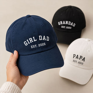 Three baseball caps are shown on a warm beige background: a navy cap held in a hand reading "GIRL DAD EST. 2026," a black cap reading "GRANDAD EST.