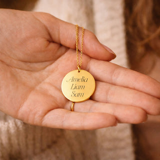 Personalised gold disc necklace engraved with family names Amelia, Liam and Sam, resting on an open hand