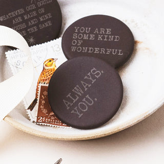 Three round dark brown engraved stone tokens or coasters displayed on a ceramic dish with vintage-style paper props. The front token reads “ALWAYS.