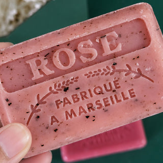 A close-up of a pink rose-colored handmade soap bar embossed with 'ROSE FABRIQUE A MARSEILLE', held by a hand against a dark green background.