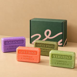 Four rectangular artisanal soaps in purple, orange, olive green, and light green are arranged in front of a dark green gift box with pink script on a beige surface and background. The soaps are labeled in French with scents including Lavande, Fleur d'Oranger, A L'Huile d'Olive, and Verveine.